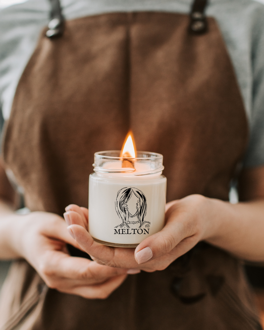 Person holding a lit candle with a visible brand logo, wearing a brown apron.